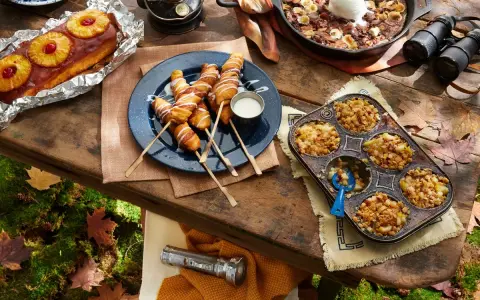 A spread of cozy camping food: pineapple upside-down cake, pigs in a blanket, apple crumble muffins, and a skillet brownie with ice cream.