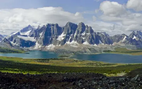 A vast mountain range with snow-capped peaks overlooks a serene blue lake. The Tonquin Valley Trail offers this breathtaking view of rugged beauty.