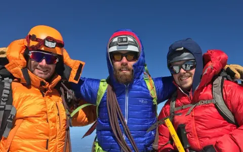 Three smiling mountaineers in bright down fill jackets and helmets pose on a sunny summit, ready for adventure.