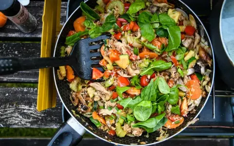 A colorful stir-fry with chicken, carrots, zucchini, and peppers, perfect for camp stove recipes. A black spatula is mixing the ingredients in a pan.
