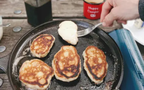 Frying pancakes in a cast iron skillet is a delicious answer to what to eat when camping. A fork lifts a fresh pancake, ready for syrup.