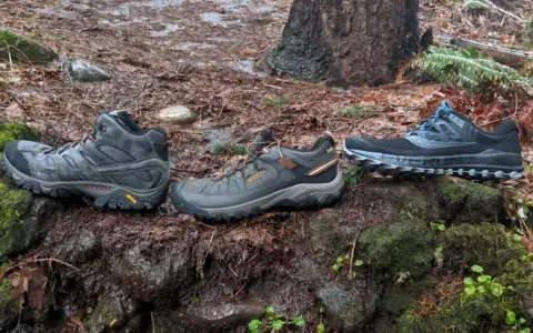 Three pairs of shoes on a wet forest floor: hiking boots, trail running shoes, and another pair of trail running shoes.