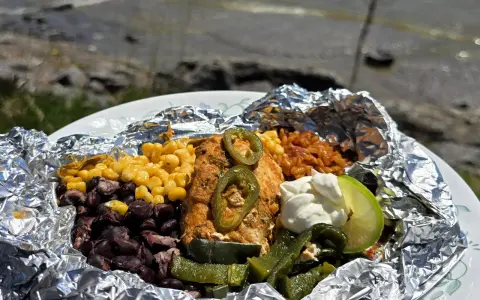 Delicious campfire chicken foil packets with corn, black beans, rice, jalapeños, and a lime wedge, ready to be enjoyed by the water.