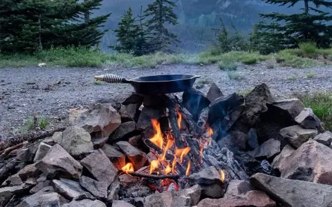 A cast iron skillet sits over a crackling campfire, ready for a delicious cast iron camping meal amidst a scenic mountain backdrop.