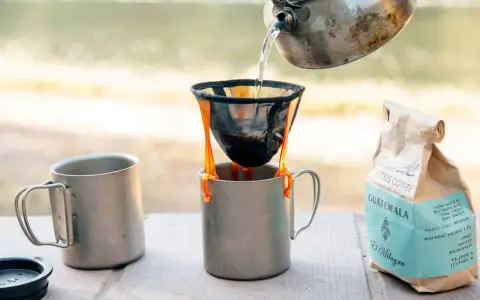Making coffee while camping: hot water pours from a kettle into a pour-over filter, brewing coffee into a metal mug. A bag of Sisters Coffee Guatemala sits nearby.