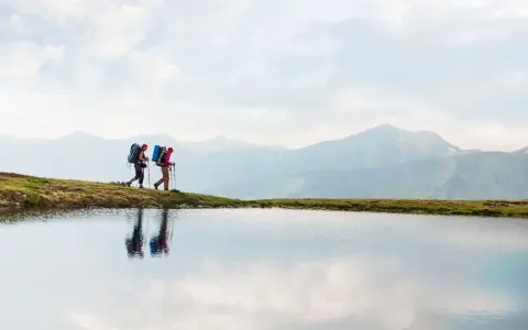 Two hikers with backpacks walk along a grassy ridge beside a calm lake, with misty mountains in the background. This image inspires thoughts on how to go backpacking.