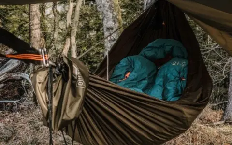 Cozy hammock camping setup in a forest. Two teal sleeping bags are nestled inside a brown hammock, ready for a night under the stars.