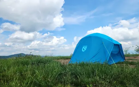 A bright blue Nemo tent stands in a grassy field under a cloudy sky. This Nemo Aurora tent is ready for adventure.