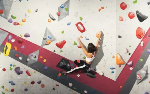 A woman is bouldering rock climbing, reaching for an orange hold on a colorful, textured wall.
