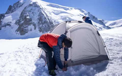 Two people set up a tent for winter camping amidst a snowy mountain landscape under a clear blue sky.