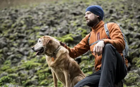 A man in an orange jacket and blue beanie rests during a hiking trip with his golden dog.