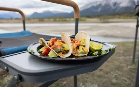 Delicious camping tacos with avocado, tomatoes, and lime, served on a plate on a portable table with mountains in the background.