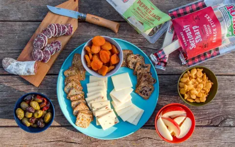 A delicious spread of camping snacks: sliced salami, cheese, crackers, dried apricots, olives, apple slices, and goldfish crackers.