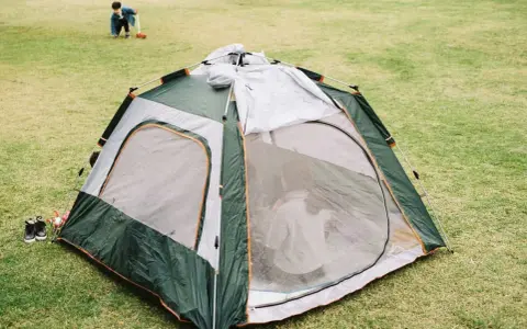 A green and gray tent is partially set up on a grassy field. A child plays in the background, learning how to set up a tent.