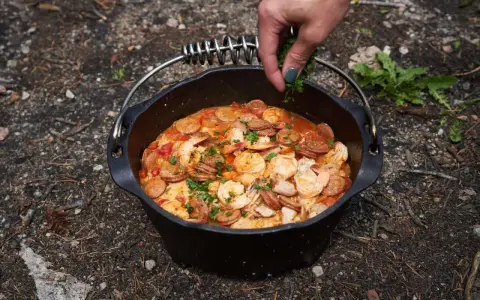 A hand garnishes a hearty dutch oven paella with fresh parsley. This campfire meal features shrimp, sausage, and rice in a rich tomato sauce.