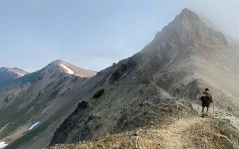 A hiker with a backpack and trekking poles walks along a narrow mountain ridge, a scenic path to learn how to get better at hiking.