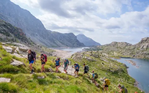 Hikers trek through a scenic mountain landscape, enjoying hiking the Pyrenees Spain. A chain of lakes and rugged peaks stretch into the distance under a cloudy sky.