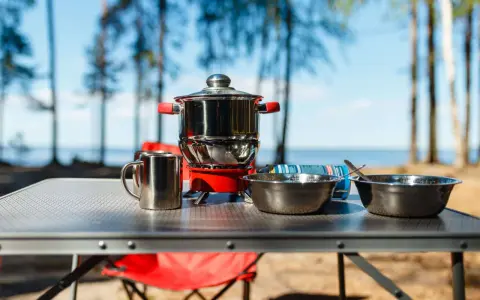 A complete camping kitchen setup with a stove, pots, bowls, and mugs on a table, ready for outdoor cooking by a lake.