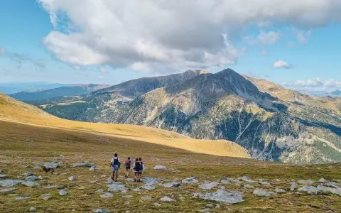 Hikers with backpacks traverse a rocky, grassy meadow on a pyrenees hut to hut hiking adventure, with majestic mountains under a cloudy sky.