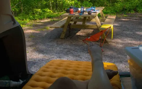 A person's legs rest on an orange sleeping pad inside a car. A picnic table with camping gear sits in a wooded area, ready for a road trip checklist.