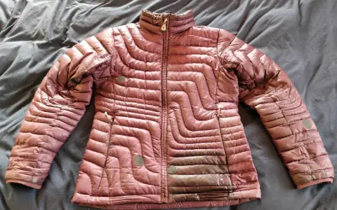 A maroon puffer jacket lies on a dark, wrinkled surface, ready for cleaning. Learn how to wash a down jacket to keep it fluffy and warm.