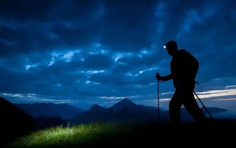 Silhouette of a hiker with a headlamp on, trekking poles in hand, on a night hike through a dramatic, cloudy sky over mountains.