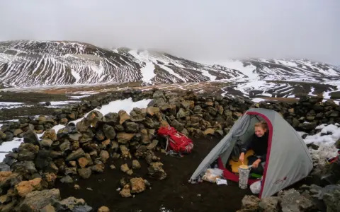 A hiker prepares a meal inside a tent at one of the Laugavegur trail huts, surrounded by snow-dusted mountains and a stone wall.