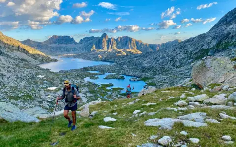 Hikers ascend a grassy slope on the HRP trail, with a stunning mountain lake and jagged peaks in the background under a cloudy sky.