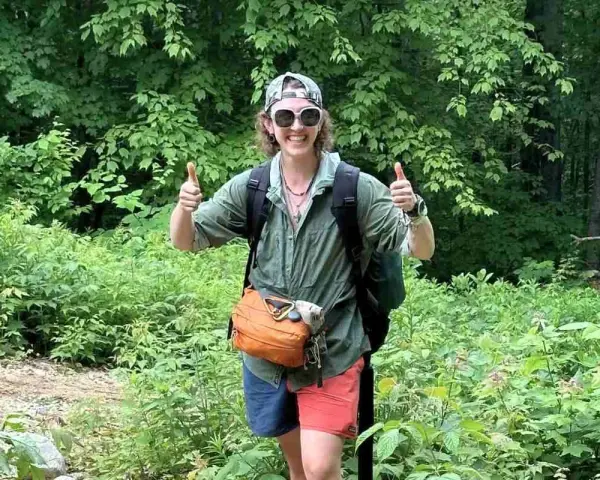 A hiker in a green, breathable hiking shirt material, gives a double thumbs-up in a lush forest.