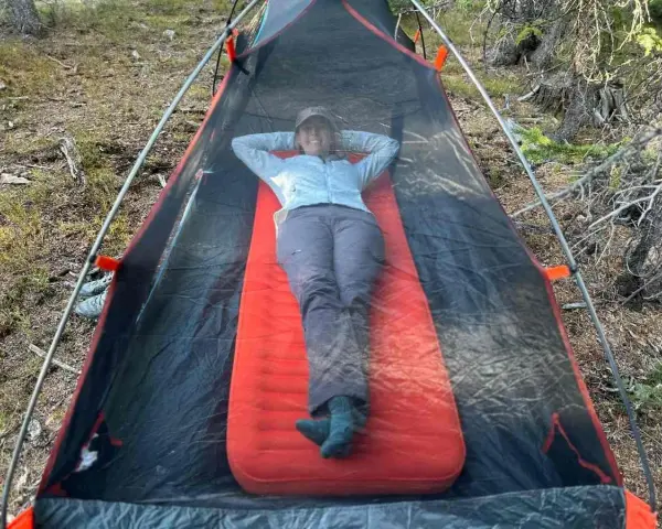 A person relaxes inside a tent on a bright orange Therm-a-Rest NeoAir XLite sleeping pad, enjoying the outdoors.