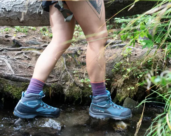 A hiker steps through a stream, testing how to waterproof hiking boots. Their blue boots and purple socks are ready for any wet trail.
