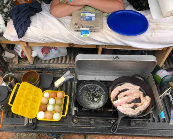 A camp kitchen checklist in action: bacon sizzles in a cast-iron pan, eggs are ready in a yellow carrier, and a portable stove is set up.