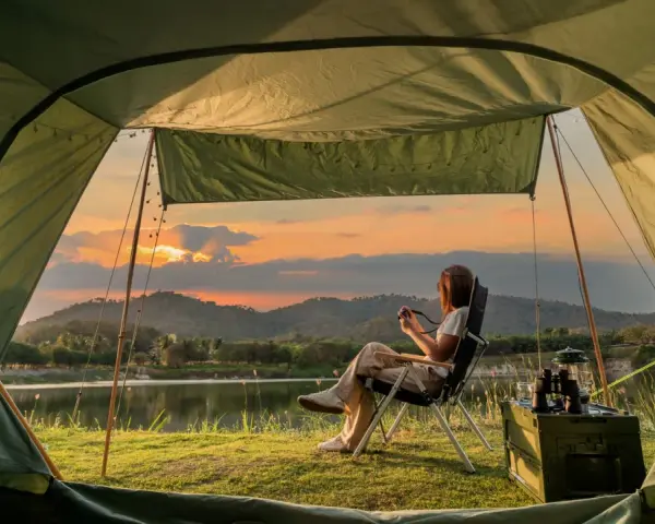 A woman enjoys solo camping by a lake at sunset, holding a camera. The tent offers a cozy view of the serene landscape.