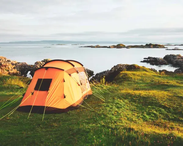 An orange tent is set up on a grassy hill overlooking a calm sea with rocky islands. This is a perfect spot for tent camp sites.