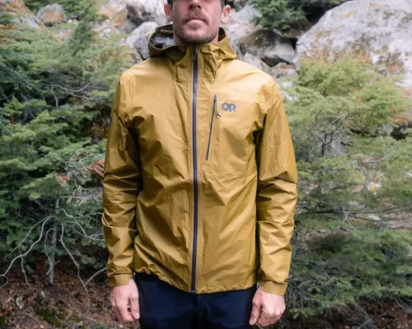 Man wearing a golden ultralight waterproof jacket with an OR logo, standing in a forest.