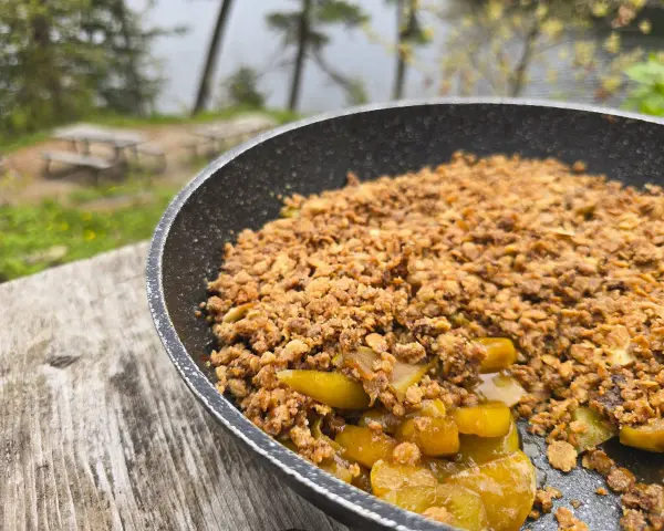 A warm, bubbling campfire apple crisp in a pan, ready to be enjoyed outdoors by a lake.