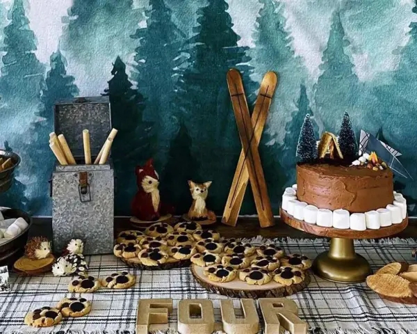 Woodland-themed camping party ideas: a chocolate cake with marshmallow border, s'mores supplies, cookies, and woodland creature decorations.