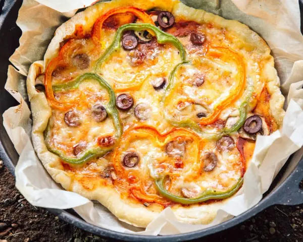 A delicious dutch oven pizza camping treat, topped with melted cheese, colorful bell peppers, and olives, baked to perfection on parchment paper.