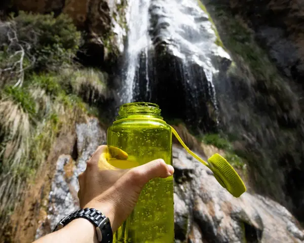A hand holds a bright green water bottle, ready for a refreshing drink after a long hike. Behind it, a beautiful waterfall cascades down a rocky cliff, surrounded by lush greenery.