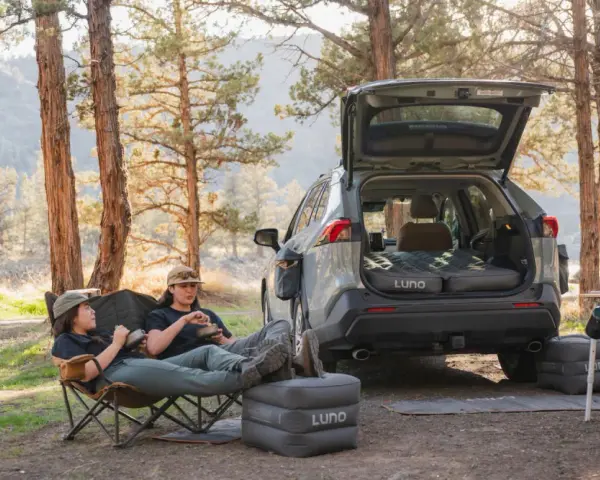 Two people enjoy a meal in camping chairs next to a car with an open trunk, showcasing clever car camping hacks with an inflatable mattress and a portable stove.