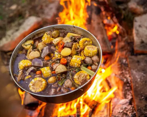A pot of hearty camping meals, filled with corn, potatoes, carrots, and meat, simmers over a crackling campfire.