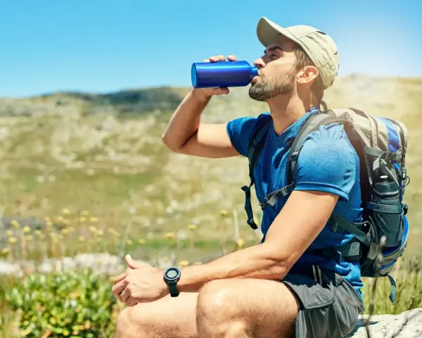 A hiker takes a refreshing drink from a blue water bottle, demonstrating how to stay hydrated while hiking in a sunny, mountainous landscape.