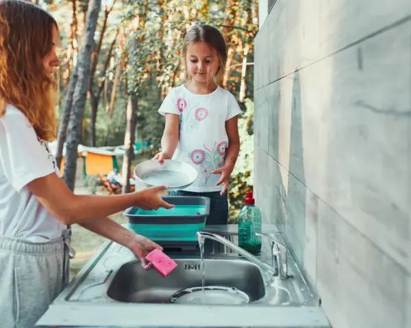 Two girls are washing dishes camping. One holds a pan over a sink while the other prepares to rinse it.