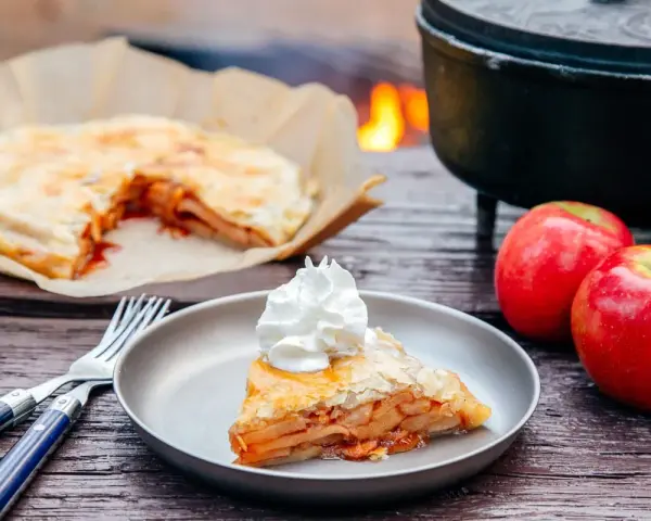 A slice of campfire apple pie, topped with whipped cream, sits on a plate next to fresh apples and a fork.