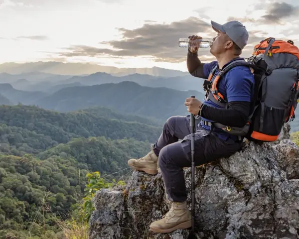 A hiker drinks water on a mountain peak, contemplating how much water to take backpacking.