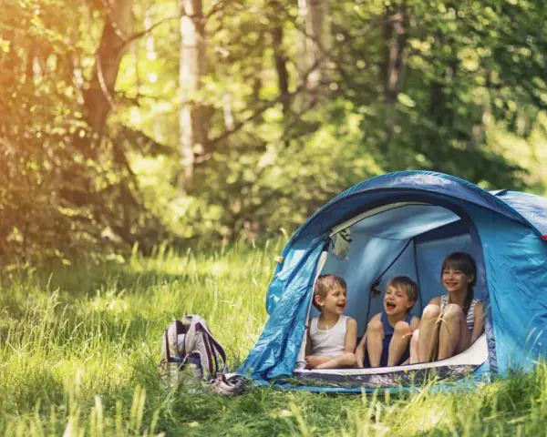 Three children laugh inside a blue tent, enjoying camping activities in a sun-dappled forest.