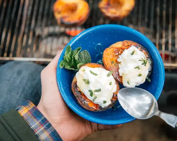 A hand holds a blue bowl with two grilled peaches topped with cream and mint. Campfire peaches are a perfect dessert.