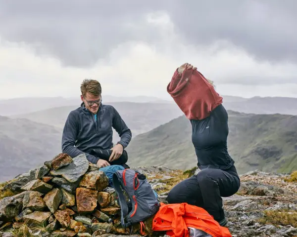 Hikers on a rocky summit adjust their backpacking clothes. One man smiles while sitting on a cairn, next to backpacks. Another person pulls on a red sweater.