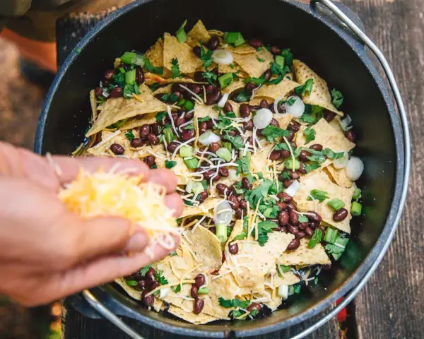 Making delicious camping nachos in a Dutch oven, adding shredded cheese to tortilla chips, black beans, and green onions.