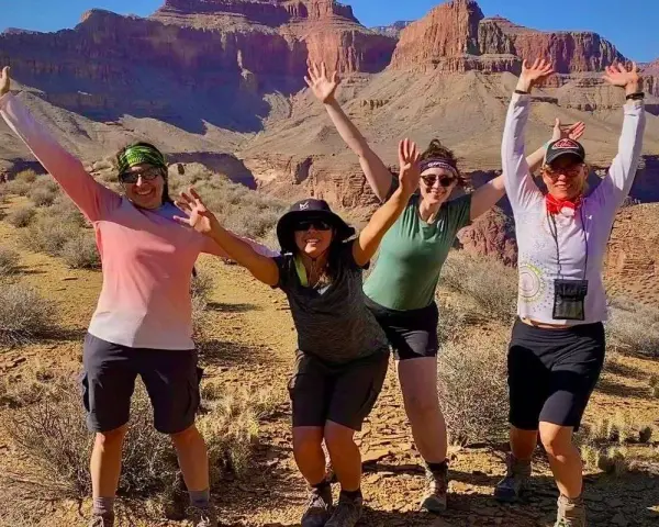Four women pose with arms raised in a desert canyon, dressed for hiking in hot weather with shorts and light tops.